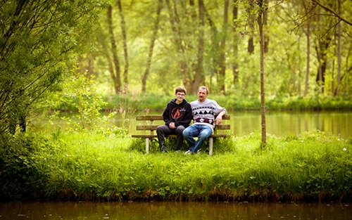 Ein Vater und sein jugendlicher Sohn sitzen entspannt auf einer Holzbank am Ufer eines Sees, umgeben von grüner Natur. Ein authentisches Outdoor-Porträt, ideal für Vater-Sohn-Fotoshootings und Familienfotos in Erfurt und Umgebung, von Michael Schalansky.